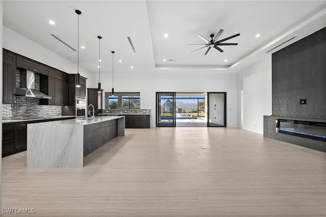 kitchen with wall chimney exhaust hood, decorative light fixtures, a tray ceiling, a large island, and backsplash