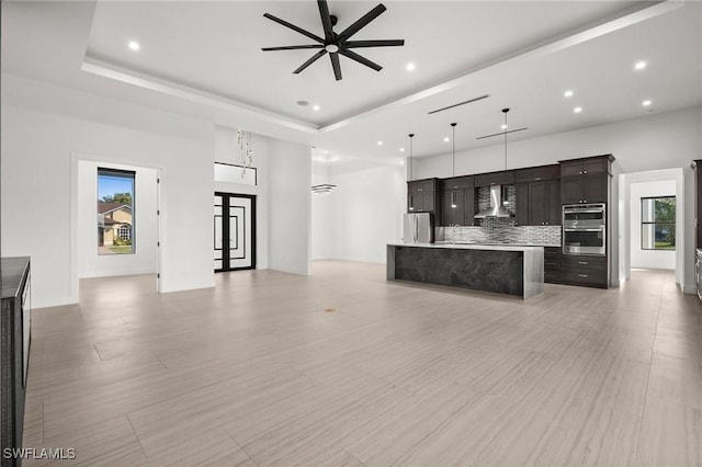 kitchen featuring wall chimney exhaust hood, decorative light fixtures, appliances with stainless steel finishes, a tray ceiling, and a kitchen island with sink