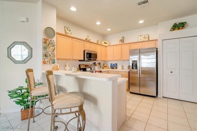 kitchen with light tile patterned floors, appliances with stainless steel finishes, a kitchen bar, kitchen peninsula, and light brown cabinets