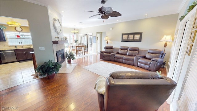 living room with sink, crown molding, light wood-type flooring, a tile fireplace, and ceiling fan with notable chandelier