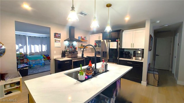 kitchen featuring pendant lighting, wall chimney range hood, appliances with stainless steel finishes, a kitchen island with sink, and white cabinets