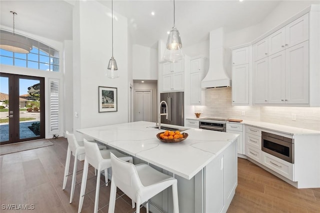 kitchen featuring a kitchen island with sink, white cabinetry, hanging light fixtures, appliances with stainless steel finishes, and custom exhaust hood