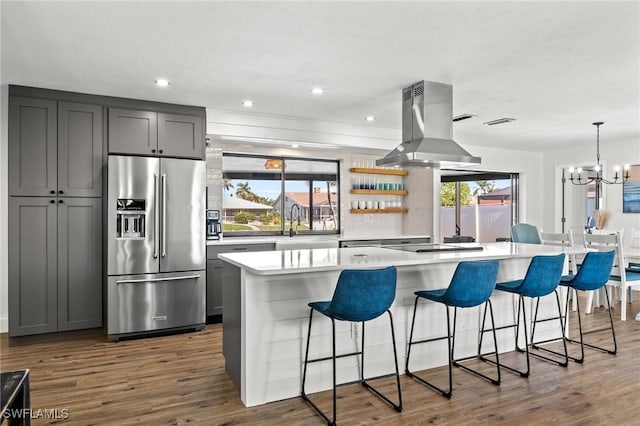 kitchen featuring a kitchen bar, high end fridge, gray cabinetry, island range hood, and a kitchen island with sink