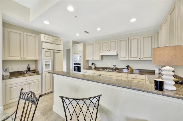 kitchen featuring built in appliances, cream cabinetry, and dark stone counters