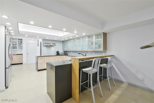 kitchen with a peninsula, wall chimney range hood, tasteful backsplash, a raised ceiling, and glass insert cabinets