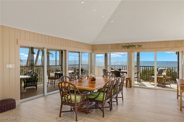 dining area featuring a water view, light wood-type flooring, wooden walls, and high vaulted ceiling