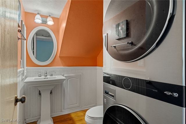 bathroom featuring stacked washer / dryer, wainscoting, toilet, wood finished floors, and a sink