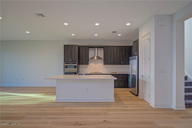 kitchen featuring wall chimney exhaust hood, dark brown cabinets, light hardwood / wood-style flooring, stainless steel appliances, and a kitchen island with sink