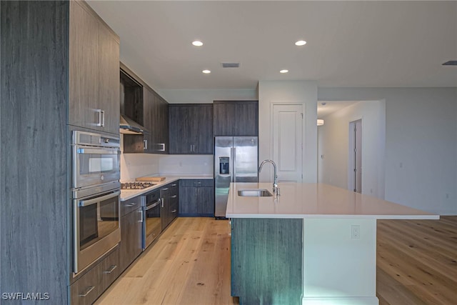 kitchen with sink, dark brown cabinets, a center island with sink, light hardwood / wood-style flooring, and appliances with stainless steel finishes