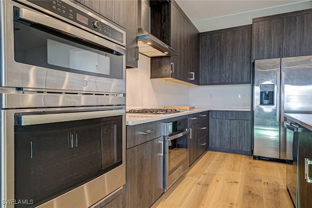 kitchen featuring stainless steel appliances, light hardwood / wood-style flooring, dark brown cabinets, and wall chimney range hood