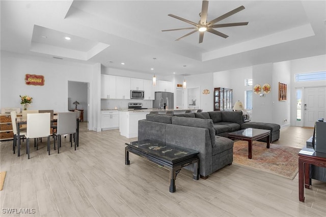 living room with a raised ceiling, ceiling fan, and light wood-type flooring