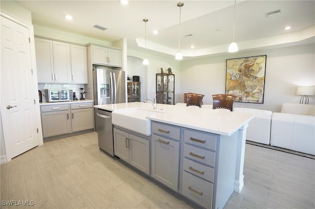 kitchen featuring sink, hanging light fixtures, appliances with stainless steel finishes, a tray ceiling, and an island with sink