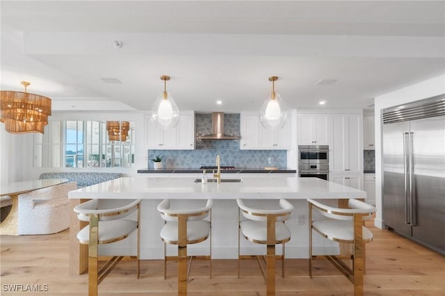 kitchen featuring pendant lighting, appliances with stainless steel finishes, white cabinetry, a kitchen island with sink, and wall chimney exhaust hood