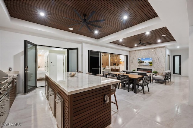 kitchen with ceiling fan, a large fireplace, a kitchen island, wooden ceiling, and a raised ceiling