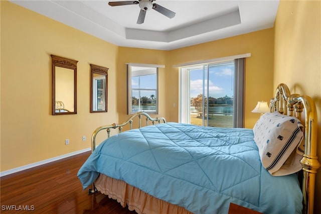 bedroom featuring dark wood-type flooring, a water view, a raised ceiling, ceiling fan, and access to exterior