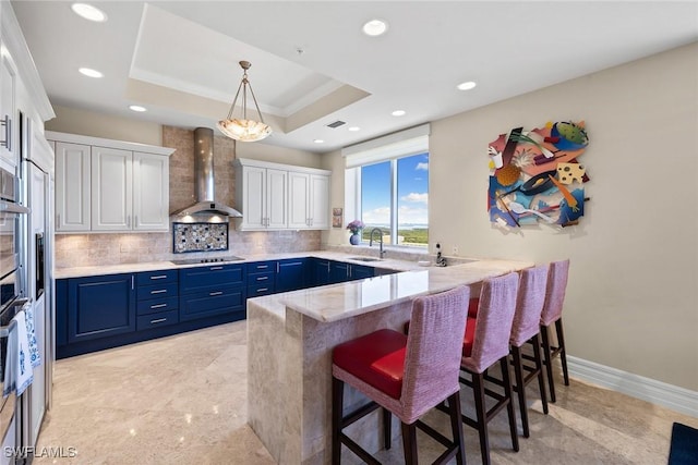 kitchen with white cabinetry, tasteful backsplash, blue cabinets, a raised ceiling, and wall chimney exhaust hood