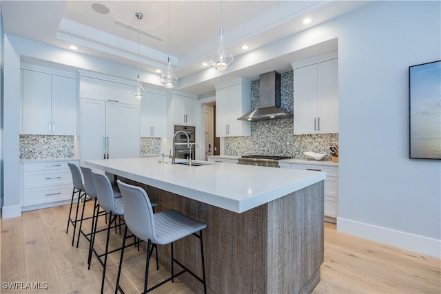 kitchen featuring a tray ceiling, light countertops, white cabinets, wall chimney range hood, and a large island with sink