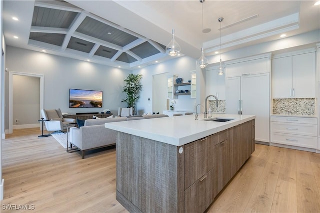 kitchen featuring sink, backsplash, coffered ceiling, white cabinets, and a center island with sink