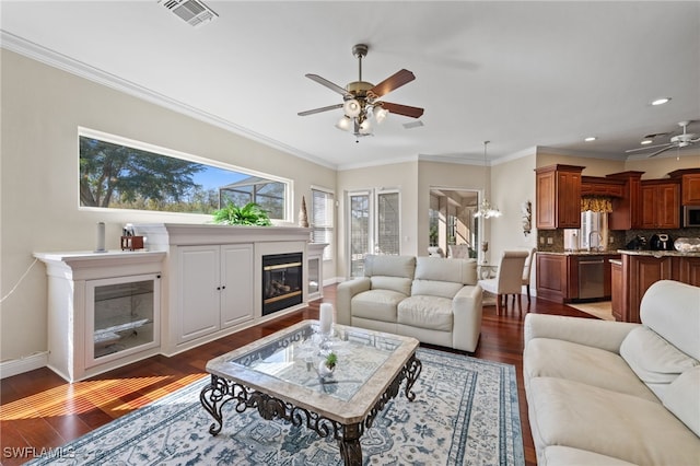 living area with ceiling fan with notable chandelier, dark wood-style flooring, visible vents, ornamental molding, and a glass covered fireplace