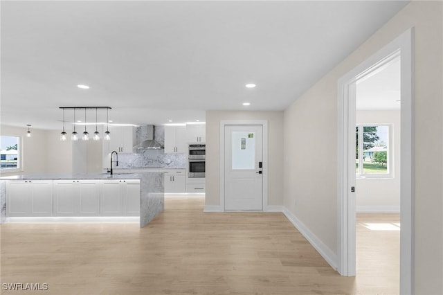 kitchen featuring tasteful backsplash, white cabinets, hanging light fixtures, wall chimney range hood, and light hardwood / wood-style flooring