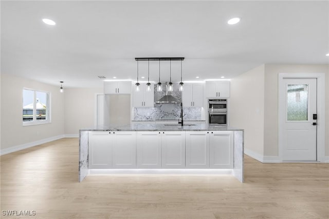 kitchen with pendant lighting, wall chimney range hood, white cabinetry, light stone counters, and stainless steel double oven