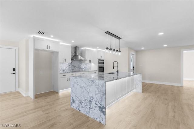 kitchen with pendant lighting, an island with sink, white cabinets, light stone counters, and wall chimney exhaust hood