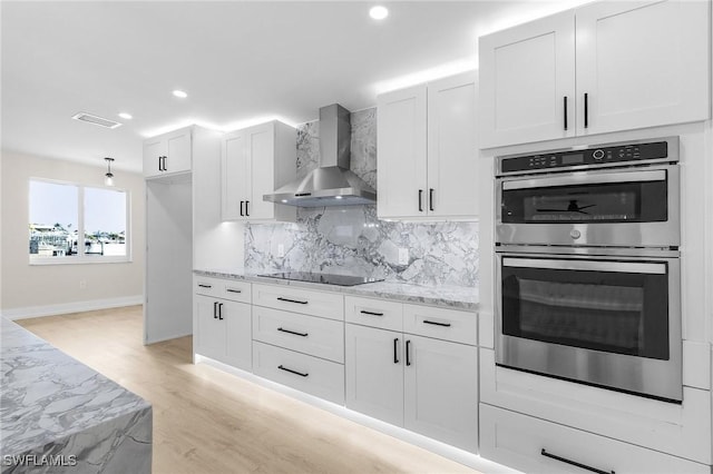 kitchen featuring double oven, wall chimney range hood, light stone counters, and white cabinets