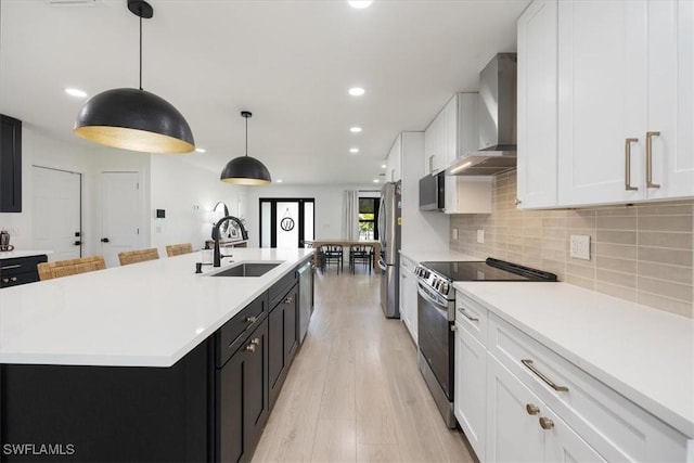 kitchen featuring wall chimney range hood, sink, appliances with stainless steel finishes, hanging light fixtures, and a center island with sink