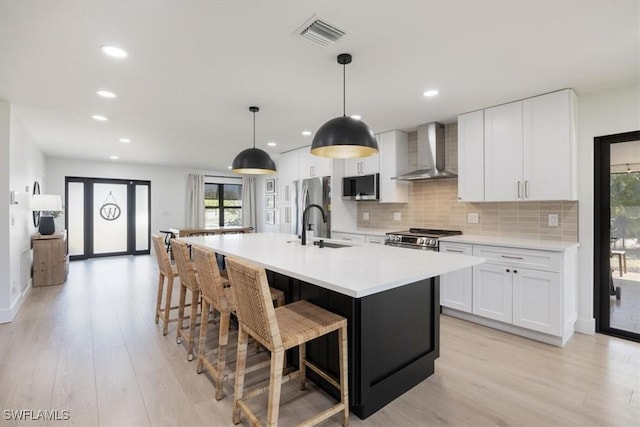 kitchen with decorative light fixtures, white cabinets, wall chimney range hood, stainless steel appliances, and a center island with sink