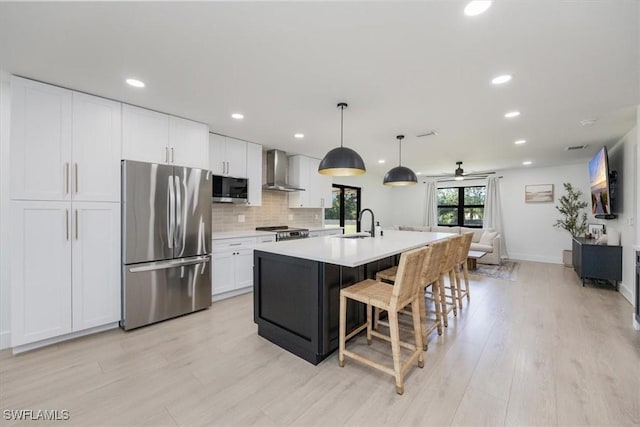 kitchen featuring decorative light fixtures, wall chimney range hood, stainless steel appliances, a kitchen island with sink, and white cabinets