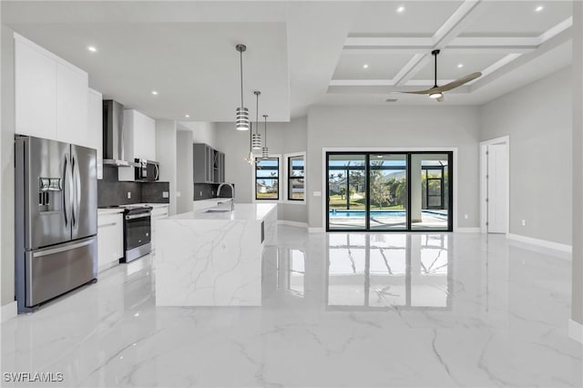 kitchen with coffered ceiling, appliances with stainless steel finishes, an island with sink, white cabinets, and wall chimney range hood