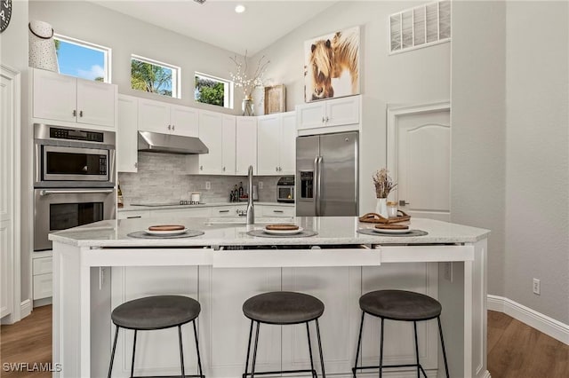 kitchen with appliances with stainless steel finishes, white cabinetry, a kitchen breakfast bar, light stone countertops, and a center island with sink