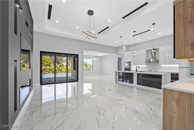 kitchen featuring white cabinetry, an inviting chandelier, decorative light fixtures, a tray ceiling, and wall chimney range hood