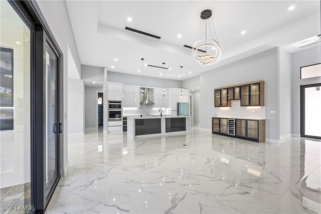 kitchen with wall chimney exhaust hood, white cabinetry, decorative light fixtures, a large island with sink, and stainless steel double oven