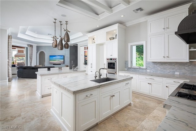 kitchen featuring wall chimney range hood, sink, light stone countertops, an island with sink, and white cabinets