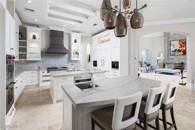 kitchen with crown molding, an island with sink, tasteful backsplash, arched walkways, and premium range hood