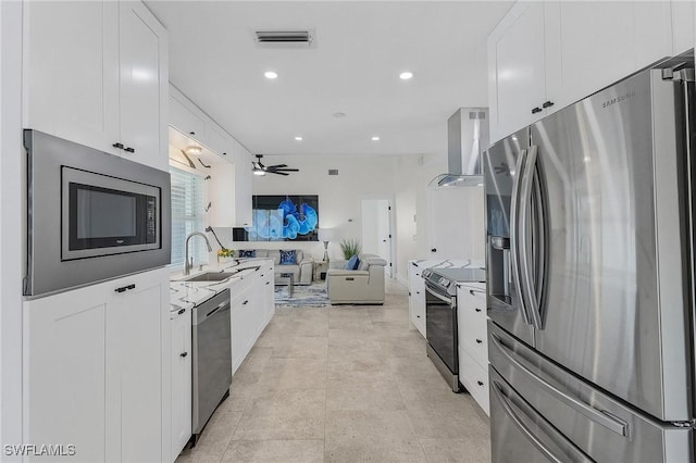 kitchen featuring wall chimney range hood, sink, ceiling fan, appliances with stainless steel finishes, and white cabinets