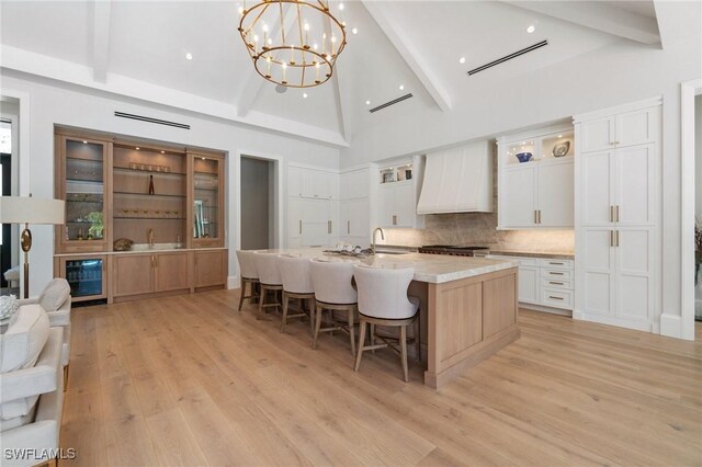 kitchen with pendant lighting, a kitchen island with sink, wine cooler, custom range hood, and white cabinets