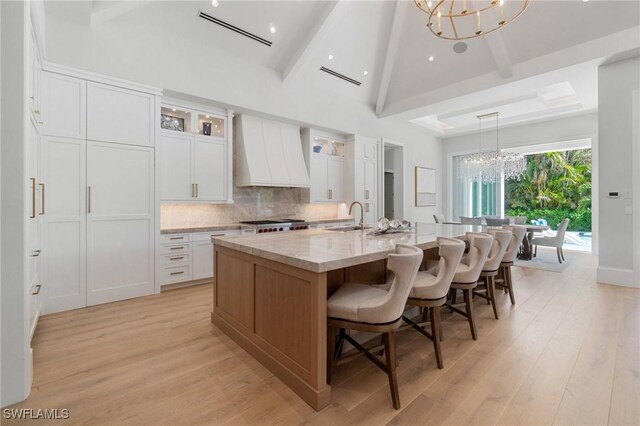 kitchen featuring sink, an inviting chandelier, custom range hood, white cabinets, and an island with sink