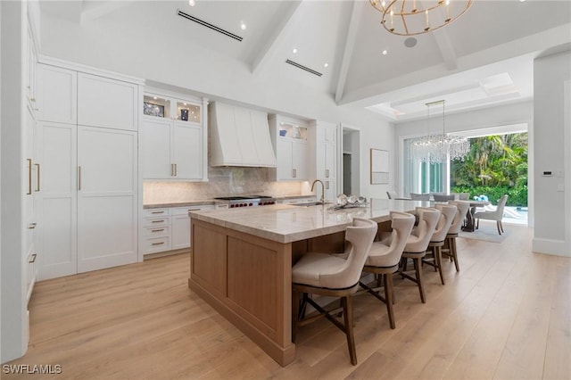 kitchen with a notable chandelier, white cabinetry, custom exhaust hood, a center island with sink, and glass insert cabinets