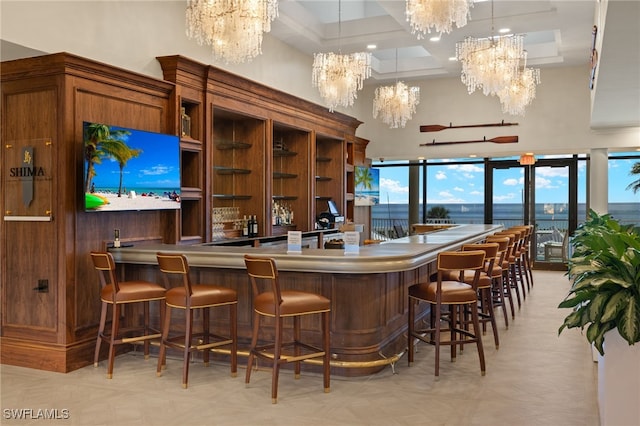 bar with an inviting chandelier, hanging light fixtures, a towering ceiling, a water view, and coffered ceiling