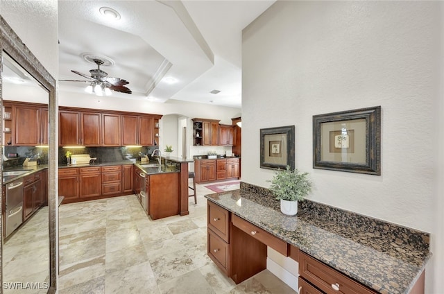 kitchen featuring ceiling fan, tasteful backsplash, built in desk, an island with sink, and a raised ceiling