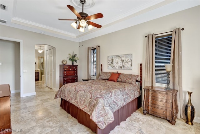 bedroom featuring ceiling fan, a tray ceiling, crown molding, ensuite bath, and a closet