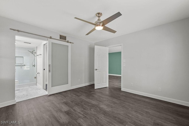 unfurnished bedroom featuring ceiling fan, a barn door, dark wood-type flooring, and ensuite bathroom