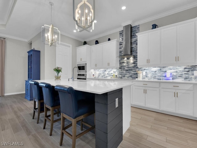 kitchen featuring white cabinetry, wall chimney range hood, a notable chandelier, stainless steel appliances, and a center island with sink