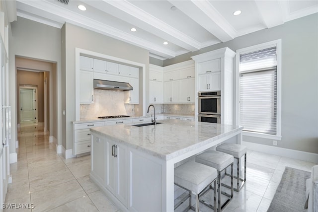 kitchen with double oven, sink, white cabinets, a kitchen island with sink, and light stone countertops