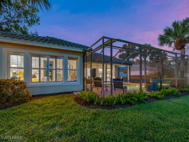 back house at dusk featuring a patio, glass enclosure, and a lawn