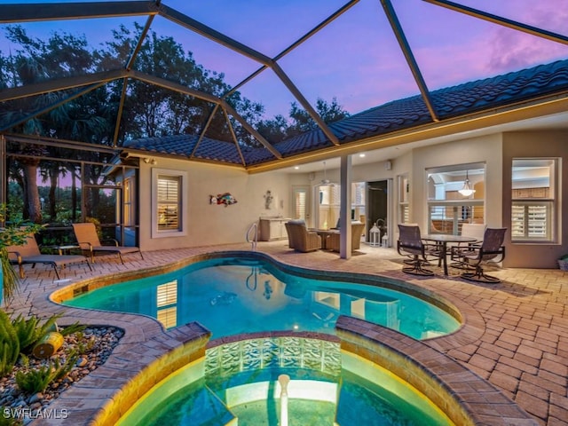 pool at dusk with an in ground hot tub, glass enclosure, and a patio