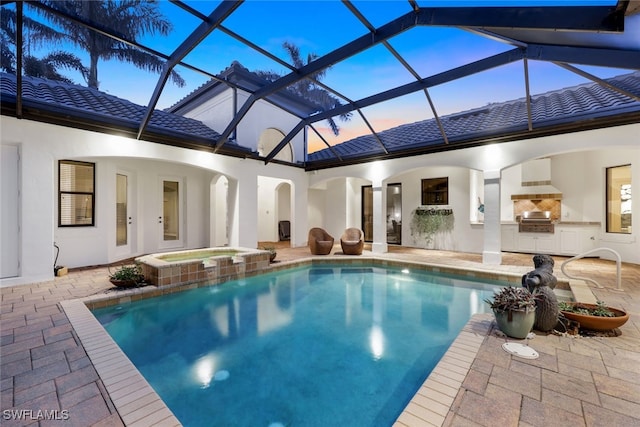 pool at dusk with a patio, glass enclosure, an outdoor kitchen, grilling area, and an in ground hot tub