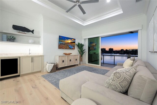 living room featuring ornamental molding, wet bar, a tray ceiling, ceiling fan, and light hardwood / wood-style floors